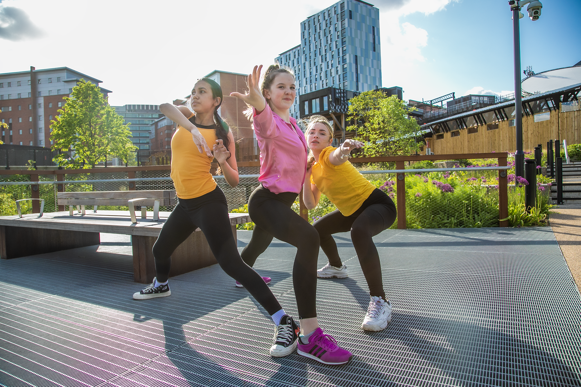 3 young female dancers infront of Machester skyline reaching out towards the camera. two girls wearing yellow tops black leggings and one girl wearing pink top black leggings 