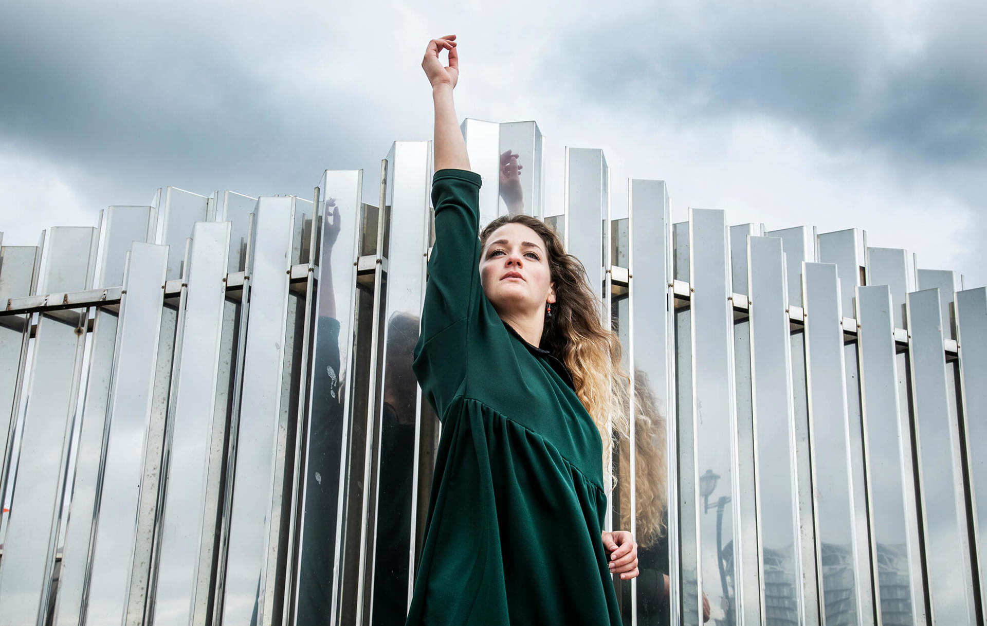 dancer standing infront of mirrored wall with stormy sky with one hand in the air. White female with curly long blonde hair and green dress 