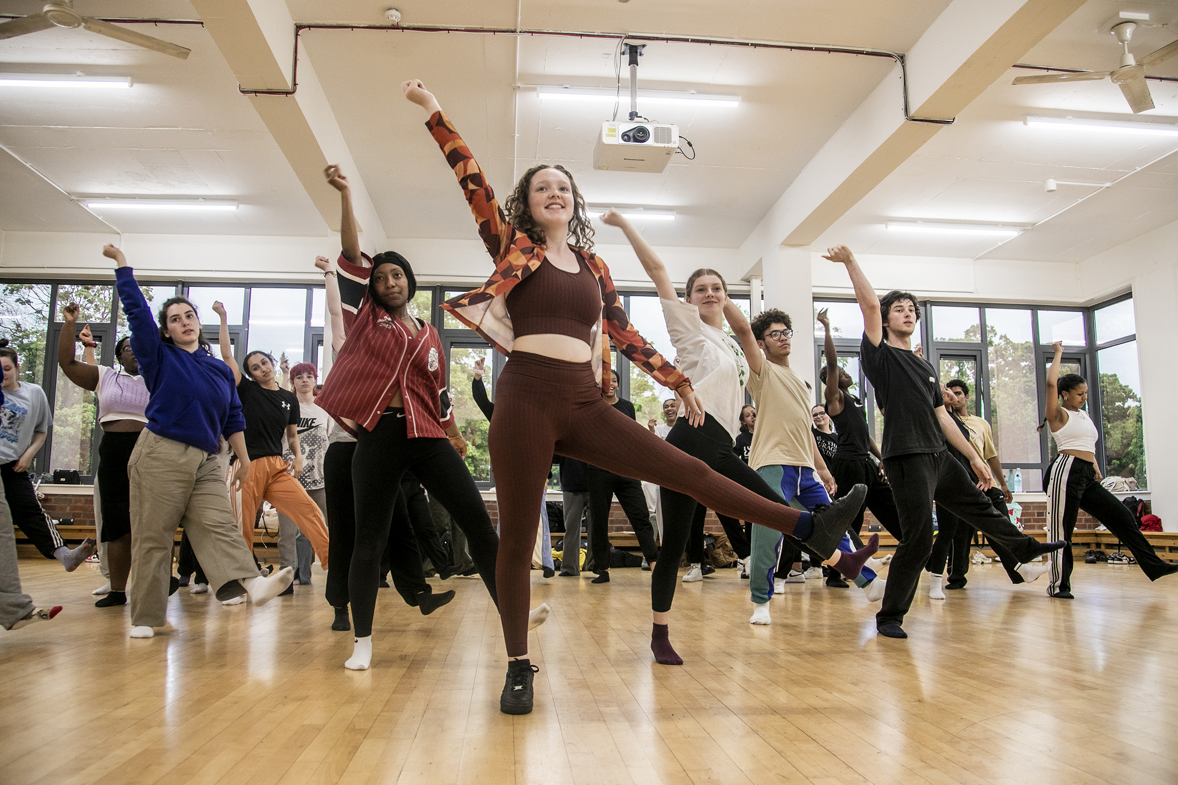 young dancers rehearing in dance studio. All with one one leg up and one arm straight leaning to the left. Mix of male and female dancers with different ethnicities. Wearing comfy casual clothes