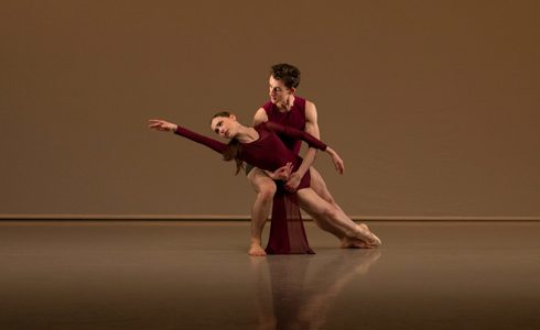 White male ballet dancer holding white female ballet dancer who is reaching to the upper left hand corner. Wearing red leotards. On a beige background