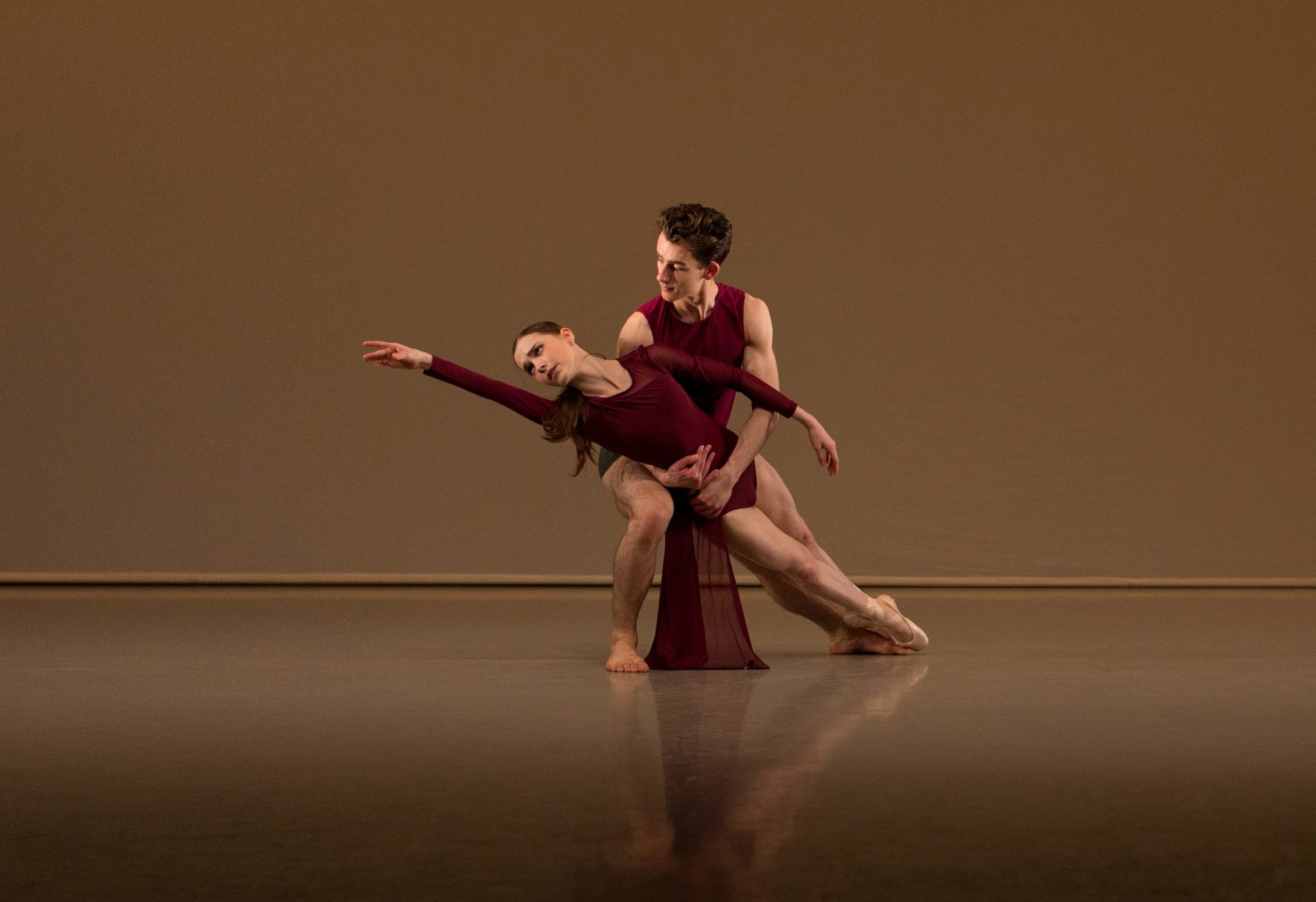 White male ballet dancer holding white female ballet dancer who is reaching to the upper left hand corner. Wearing red leotards. On a beige background