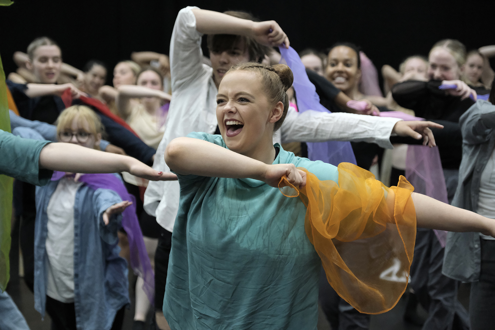 young white female dancer with red hair in a bun wearing blue top with a big happy face, holding an orange scarf with arms to the right with other young dancers behind doing the same movement. All in colourful clothes holding scarfs.