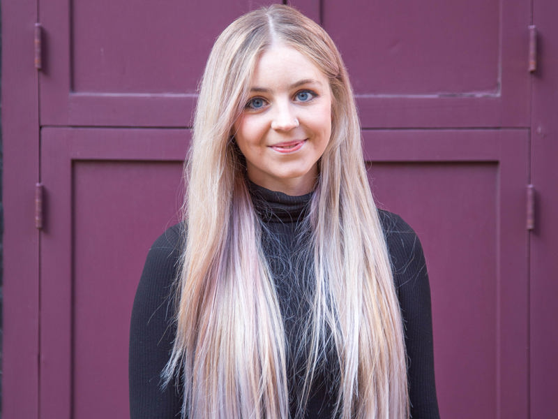 Headshot of Amelia Wilkinson. White female with long blonde hair smiling at the camera wearing long sleve black top in front red door