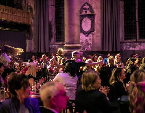 audience sitting down clapped as drag show dancers dance through the seats and tables. Large church venue is lit pink 