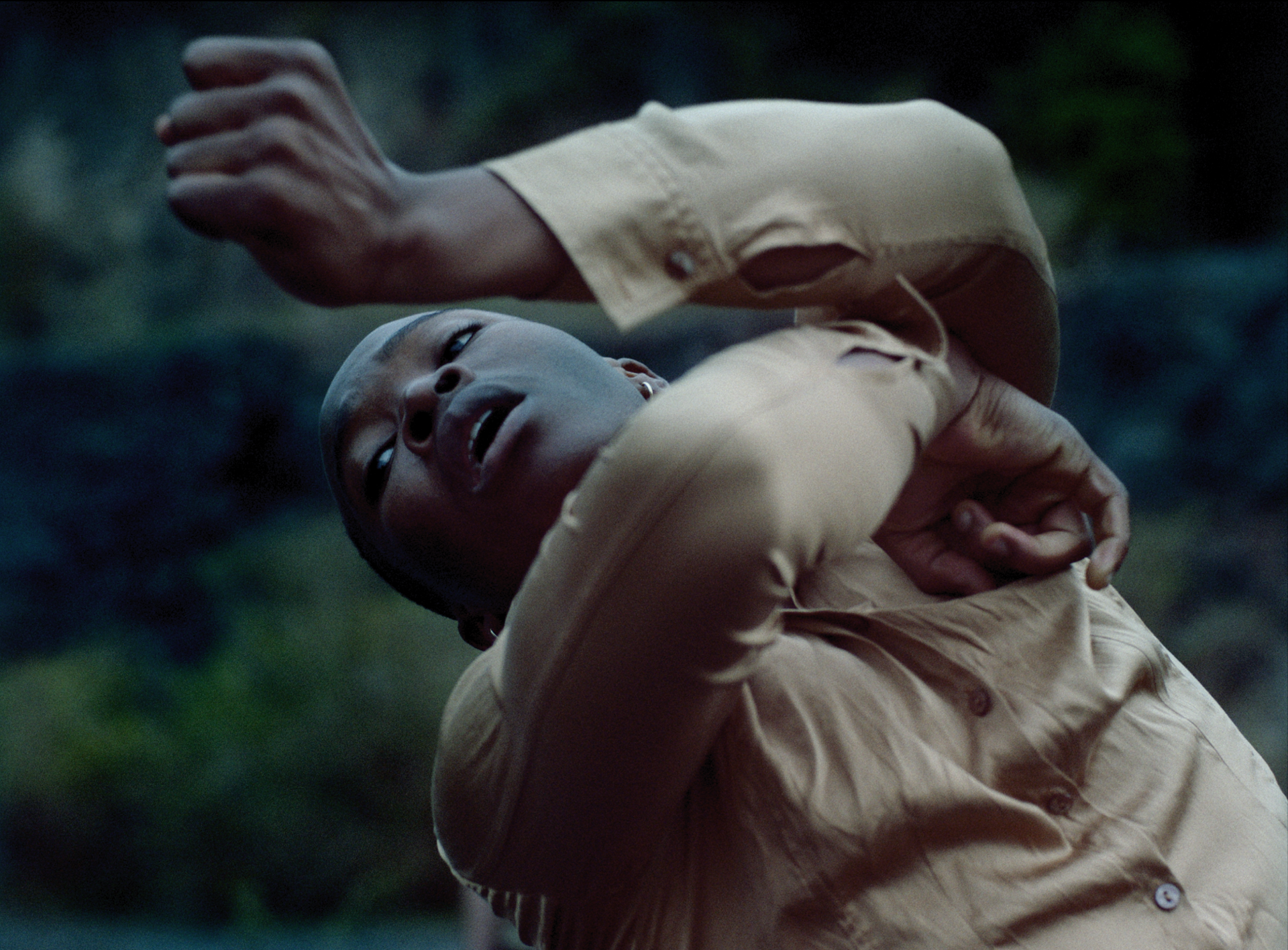 Close up of global majority dancer moving arms around the face with eyes up to the top left corner of the image, wearing muted clothes in front of dark wall 