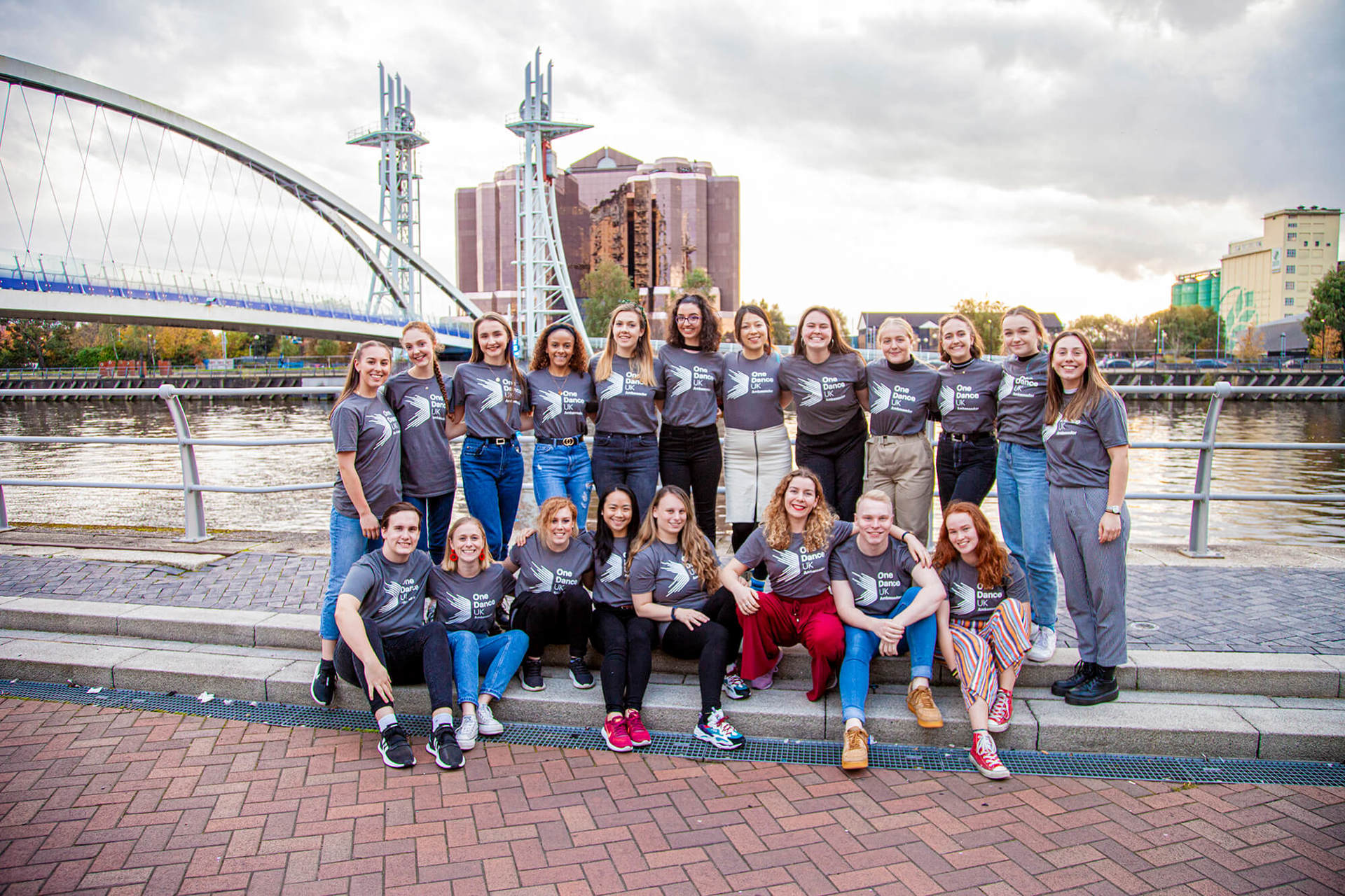 20 male and female dance ambassadors linked arms smiling in front of Manchester bridge and skyline in the background. Wearing grey Dance ambassador tshirts.