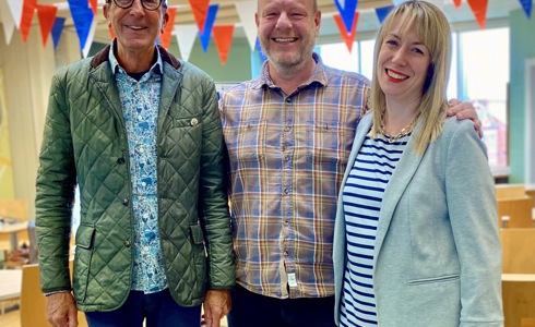 two white men and a white women standing smiling looking at the camera in a room with red blue and white bunting 