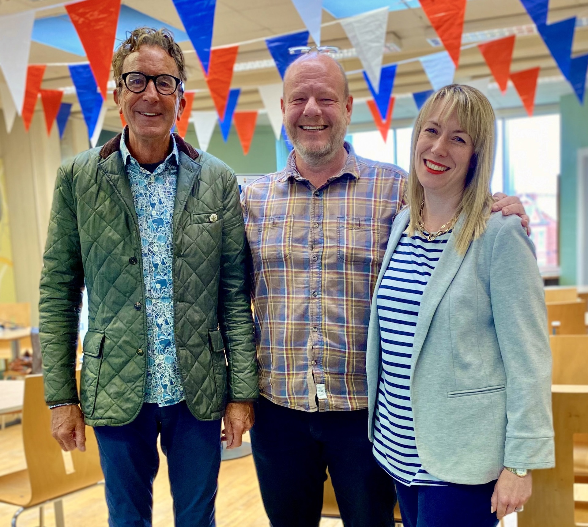 two white men and a white women standing smiling looking at the camera in a room with red blue and white bunting 