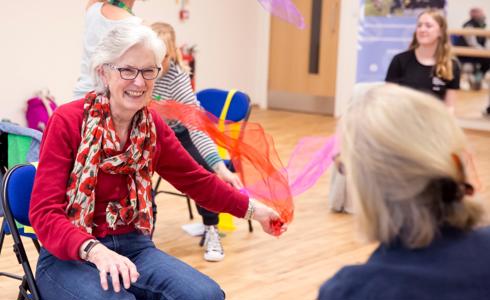 Older female sitting on a chair waving a colourful scarf around whilst smiling 