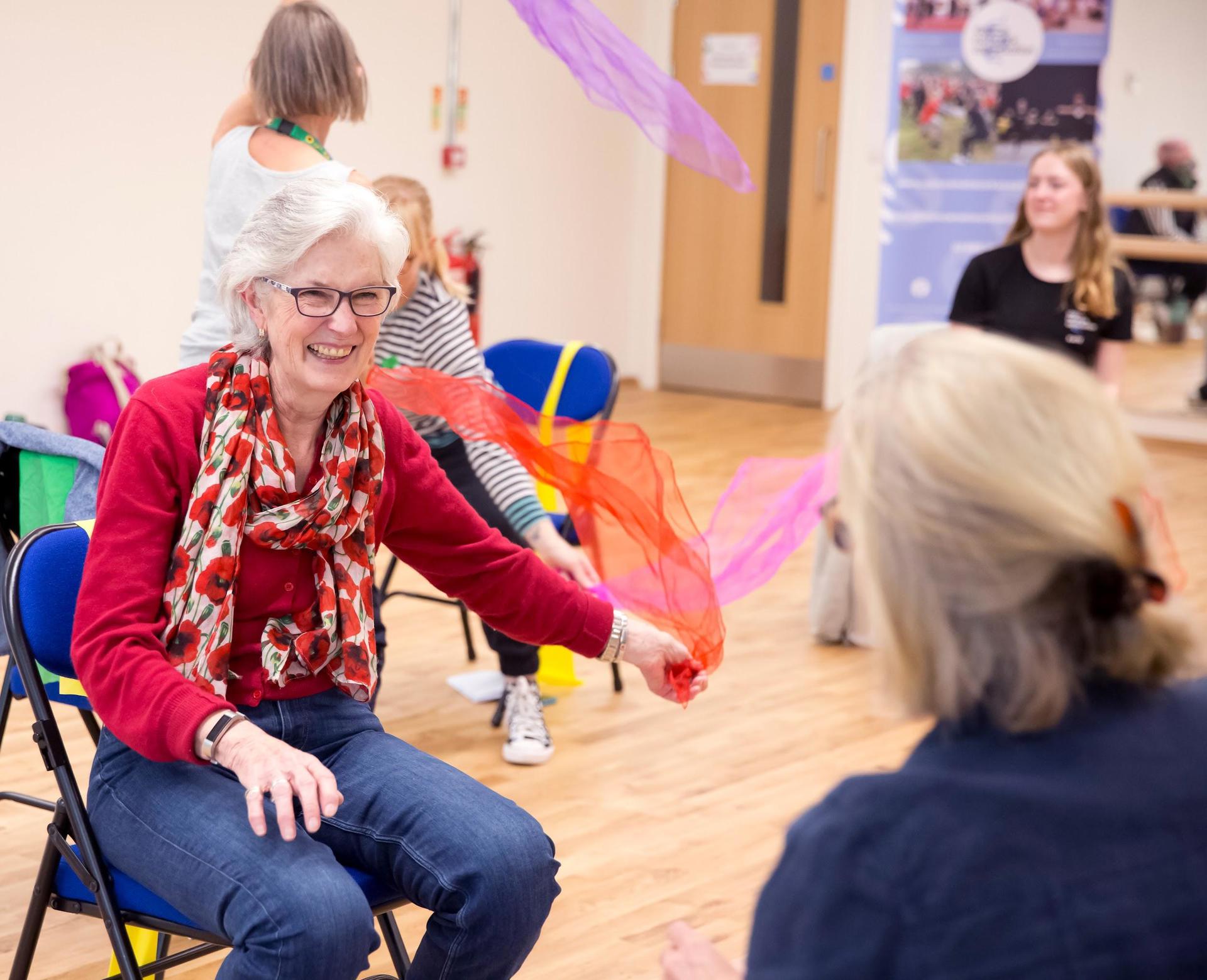 Older female sitting on a chair waving a colourful scarf around whilst smiling 