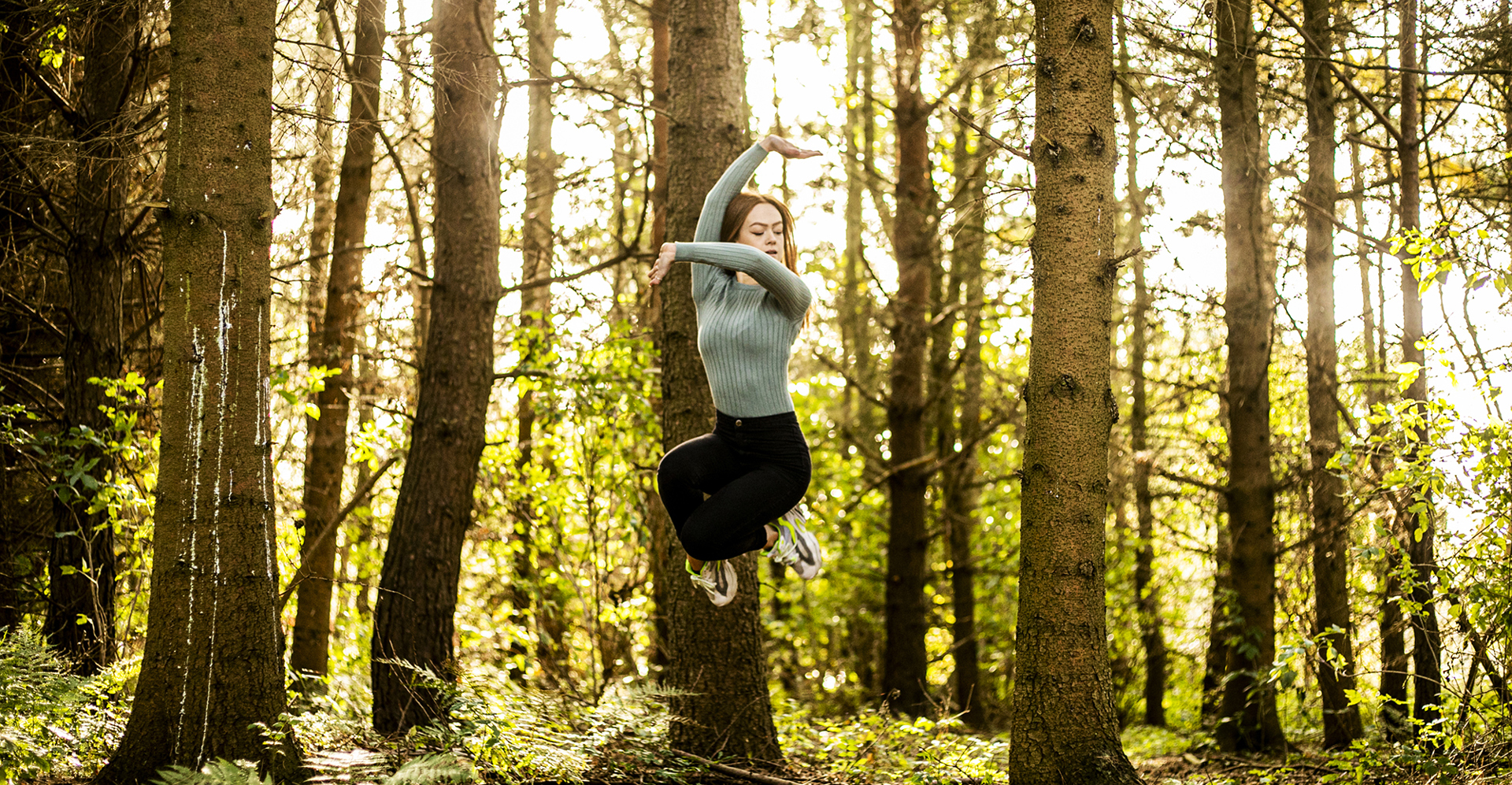 white female dancer jumping in the air with legs tucked and one arm up and the other across body. In front of woodland in golden light.