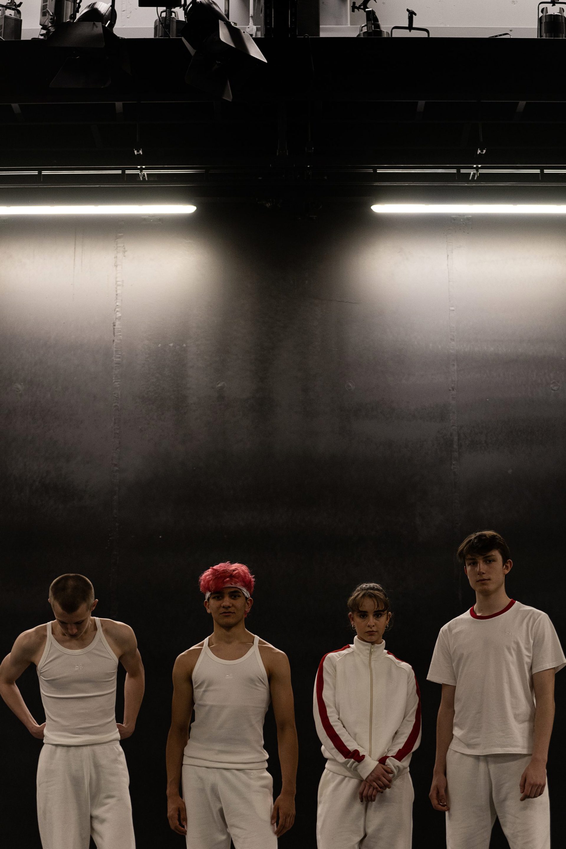 four young dancers wearing white tracksuits looking at the camera with dark walls behind them 