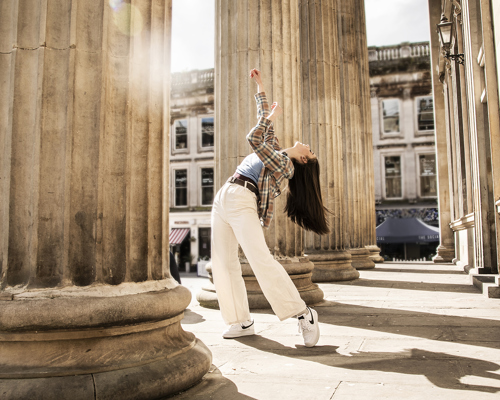 Young female dancer infront of Glasgow coucil building with large pillars. Legs lunched arms above the head. White female dance with long brown hair wearing checked shirt blue top and white trousers 