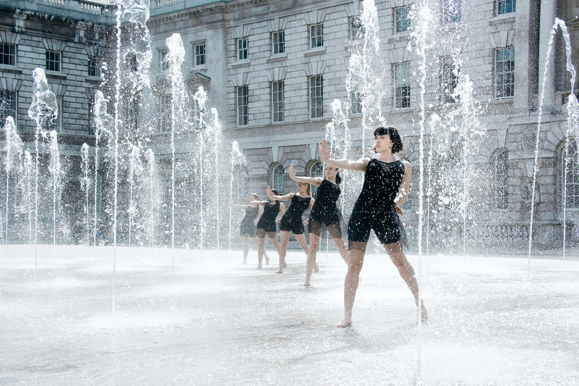 five female dancers with one arm reached out all standing in a water fountain. Wearing black outfits.