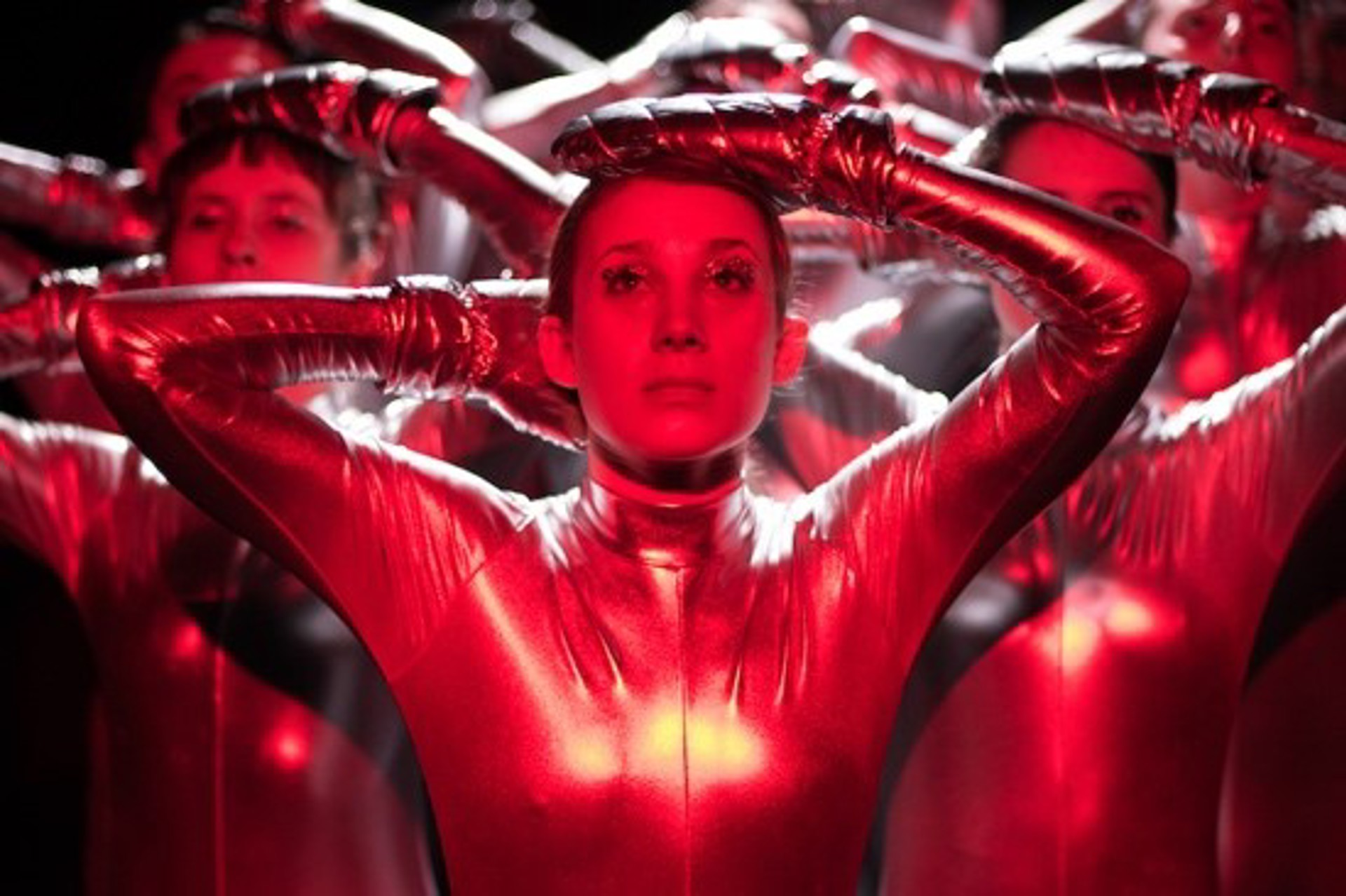 female with hands on head with other dancers in the background copying the movement. All wearing lycra bodysuit with red lighting. 