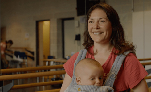 White female holding baby in a body carrier talking to the camera in a dance studio