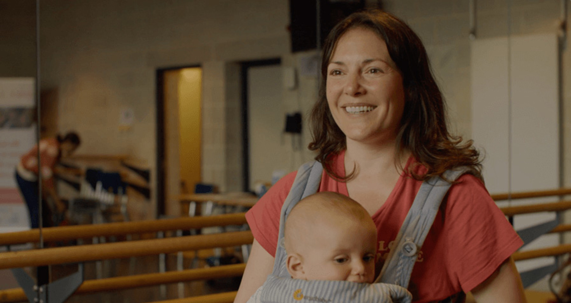 White female holding baby in a body carrier talking to the camera in a dance studio