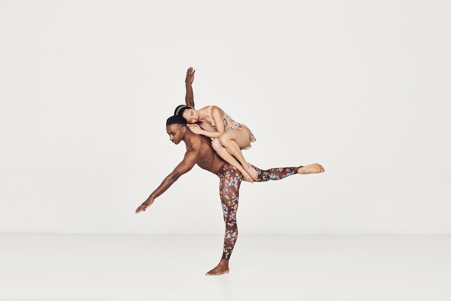 Global majority male dancer on one leg with arms stretched wise with global majority female dancer curled up balancing in his back in a white studio