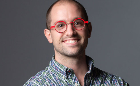 Headshot of Constant Vigier. White male with shaved head smiling at camera wearing red circle glasses and checked shirt 