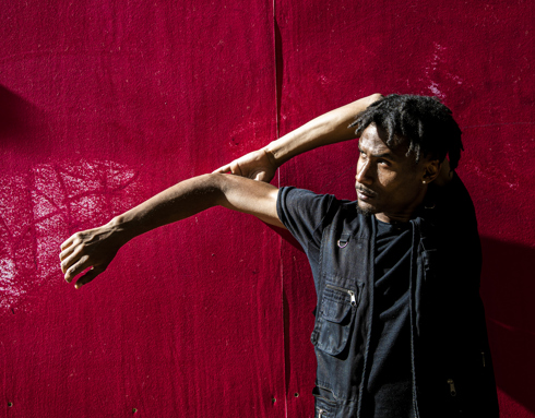 Global majority male dancer with short dreaded hair, one arm strecthed to the left and the other holding the elbow behind his head. Wearing all black standing in front of a red wall .