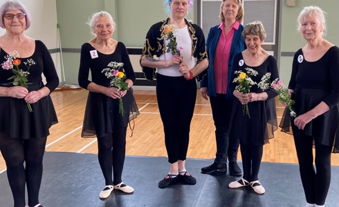 Six older white Bbodance dancers wearing black ballet costumes holding flowers and looking at the camera. In a dance studio,