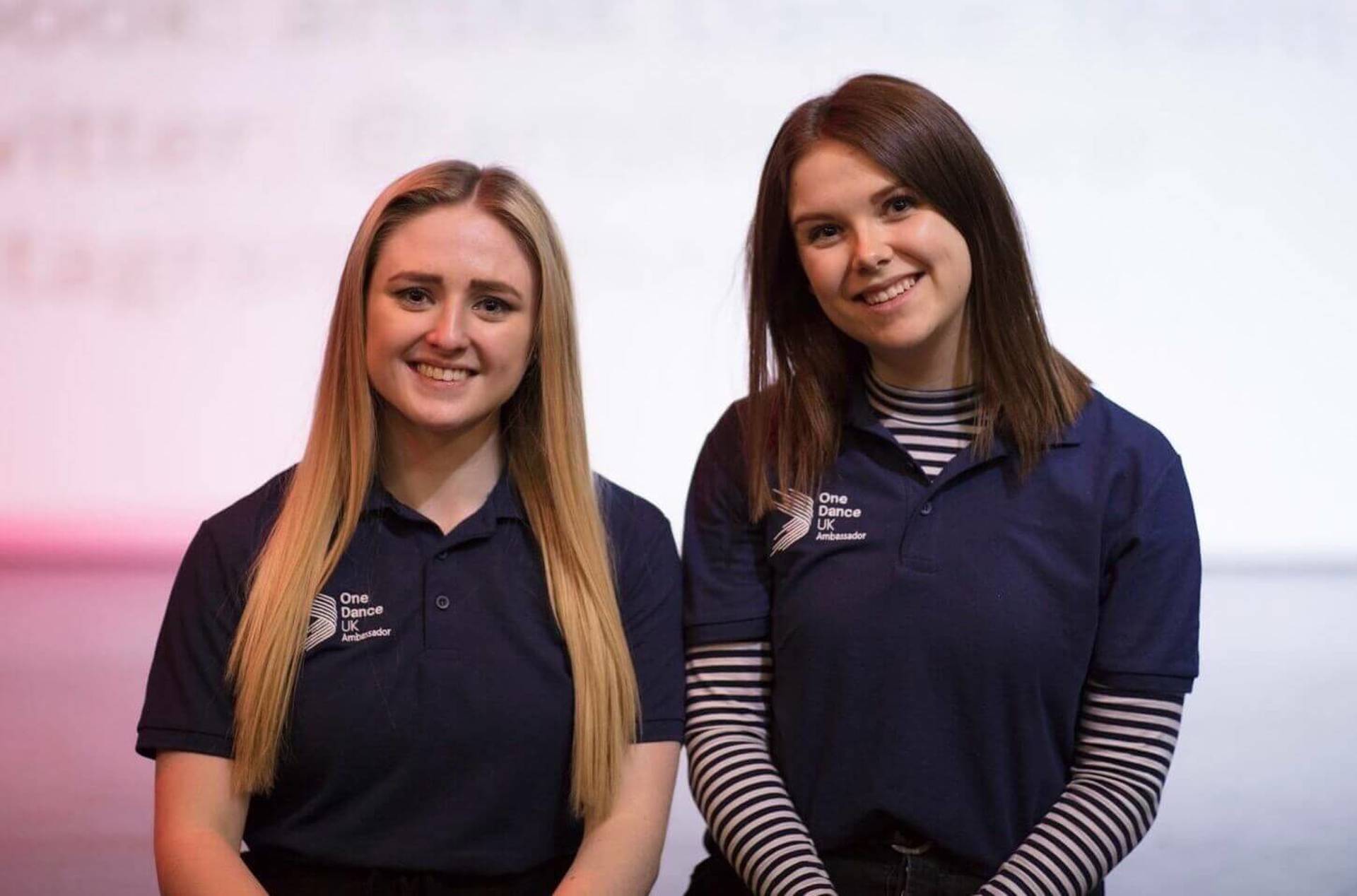 Two female One Dance UK dance ambassadors in blue t-shirts smiling at the camera