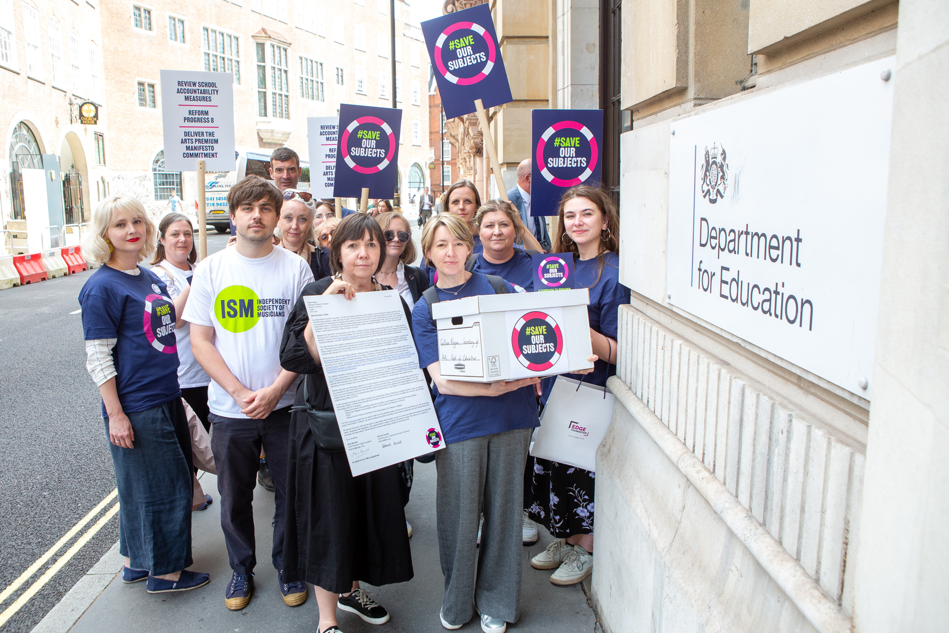 fourteen adults mixed genders standing in a group holding signs that say save our subjects outside the department of education