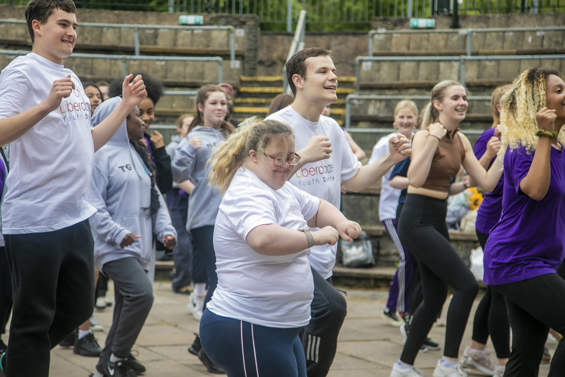 Young people dancing in an outdoor warm up, central dancer is a white female smiling. Wearing a mix of coloured outfits. 