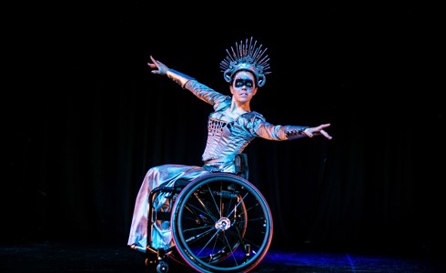 White female wheelchair dancer on stage wearing snow queen costume including large spike crown and black face paint across the eyes