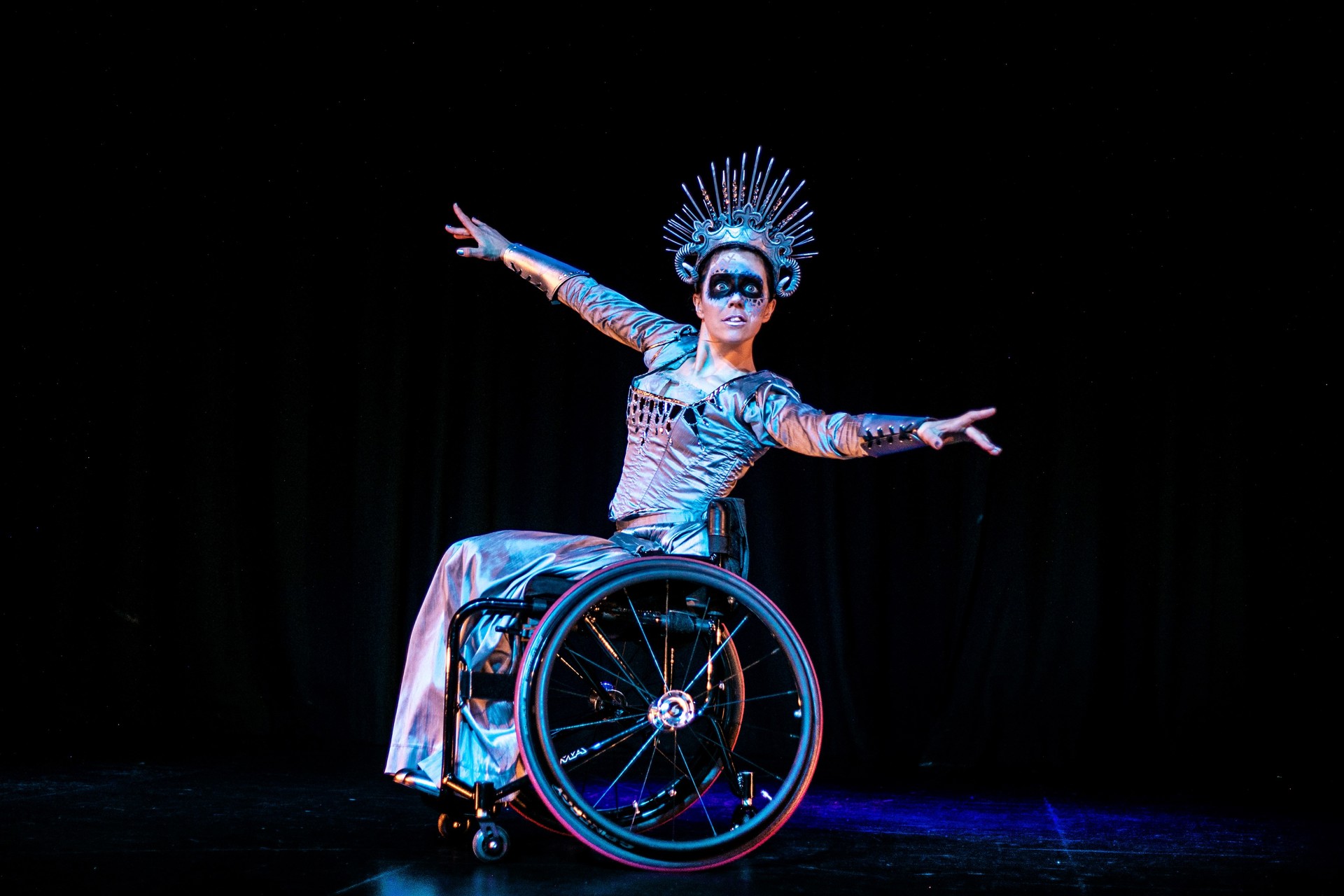 White female wheelchair dancer on stage wearing snow queen costume including large spike crown and black face paint across the eyes