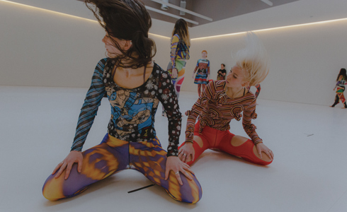 two female dancers sitting on their knees with hands on knees looking left and flicking hair. Wearing colourful patterned clothes 