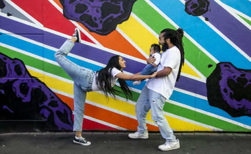 Family of 3. Global majority male with long dreadlocked hair holding baby, global majority female with long black hair, one leg up then reaching our leaning towards the baby. wearing blue jeans, trainers and white tshirts infront of rainbow graffiti wall. 