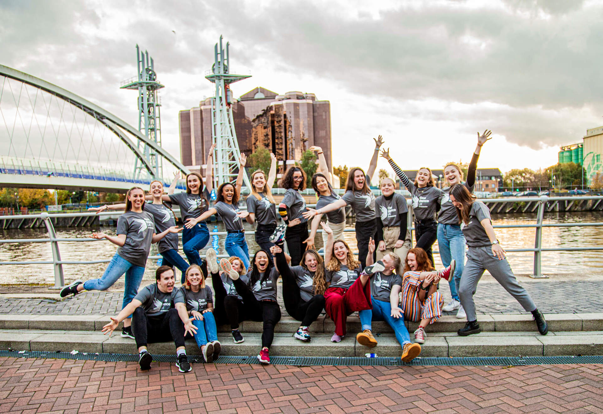 20 male and female dance ambassadors posing in front of Manchester bridge and skyline in the background. Wearing grey Dance ambassador tshirts.