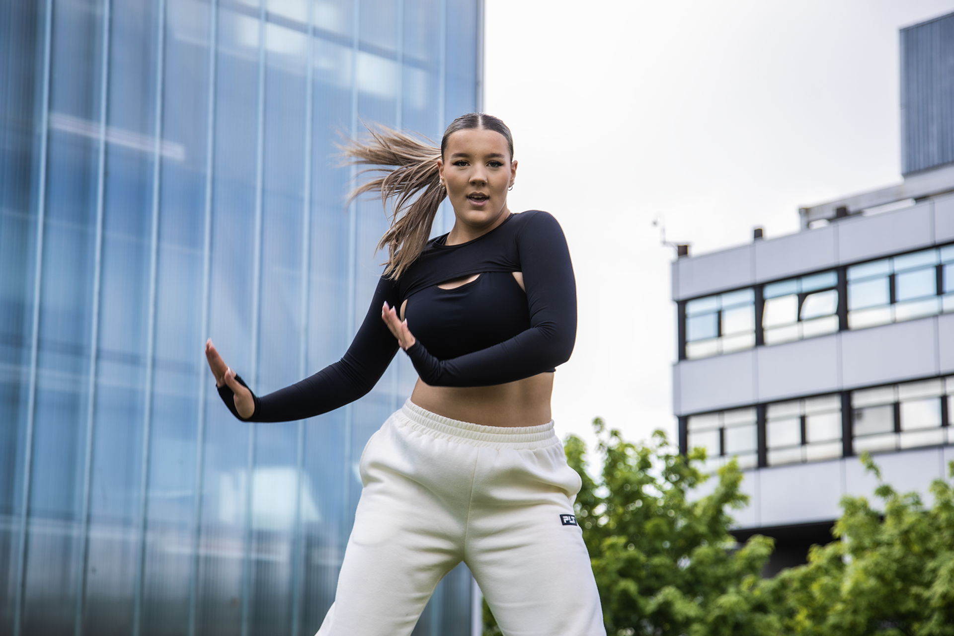 young female dancer with arms to the right side of her infront of Newcastle college building. Wearing black top, white joggers and ponytail swinging to the left