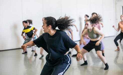 Female global majority dancer with curly hair flicked in a pony tail teaching a hip hop class. students copying the movement behind all wearing bright colours in dance studio