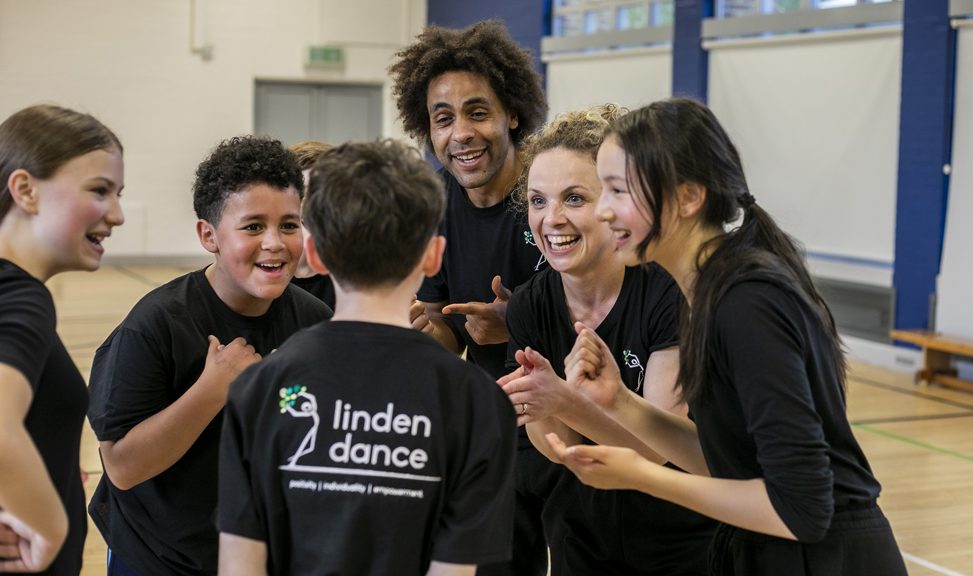 teachers and dance students excitedly talking together in black PE kit with sports hall