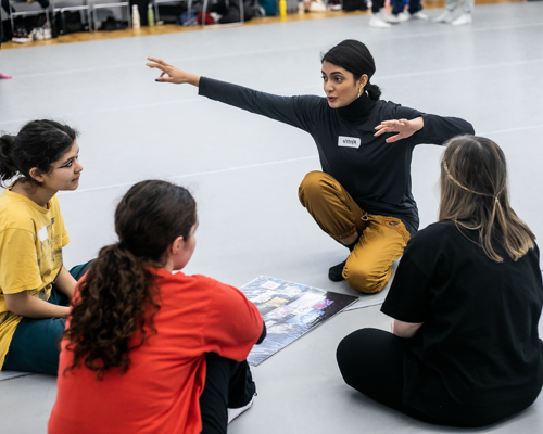 Global majority dance teaching kneeling down with 4 young female dance students gesturing with arms out wide with a mood board in front of them in a dance studio. 