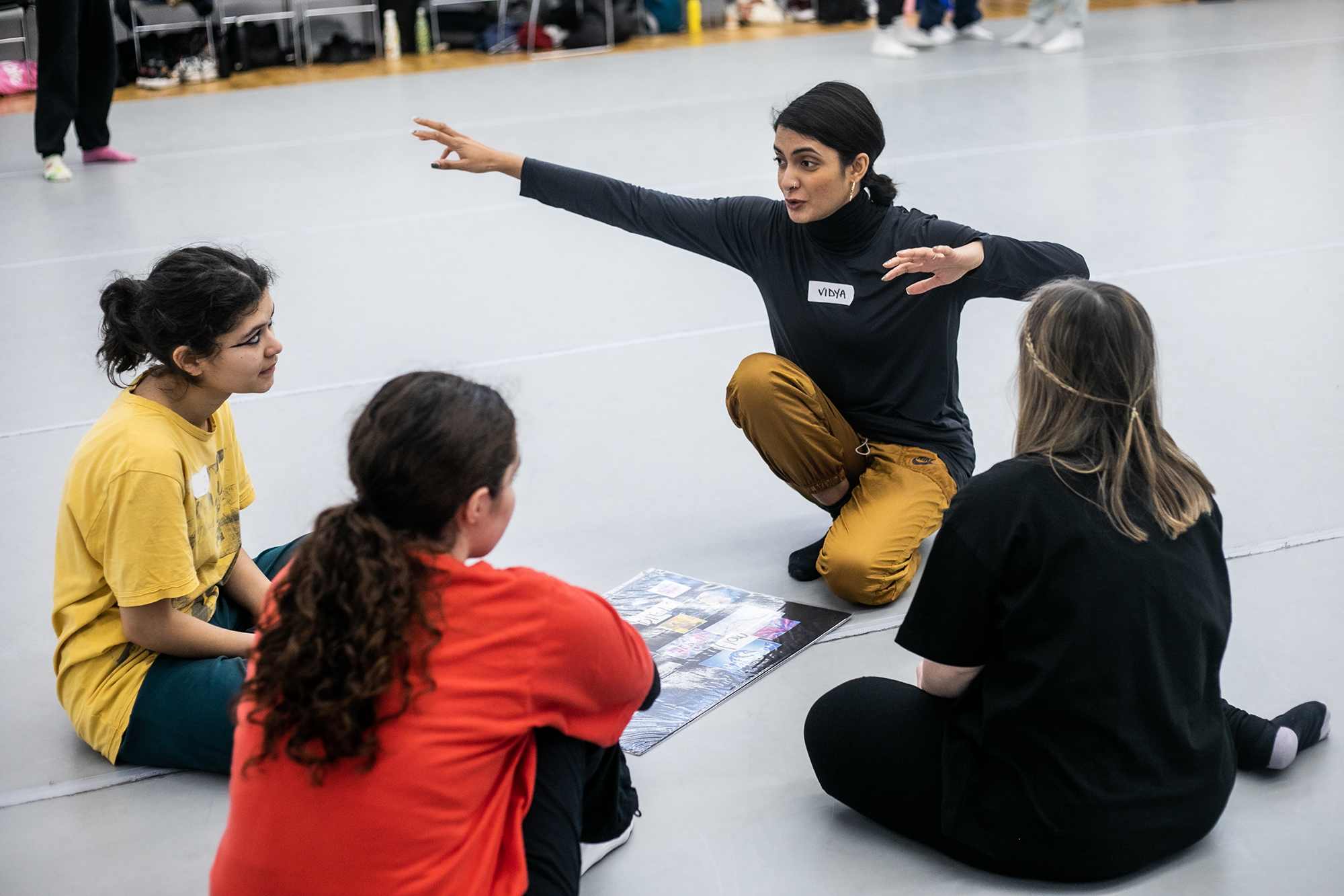 Global majority dance teaching kneeling down with 4 young female dance students gesturing with arms out wide with a mood board in front of them in a dance studio. 