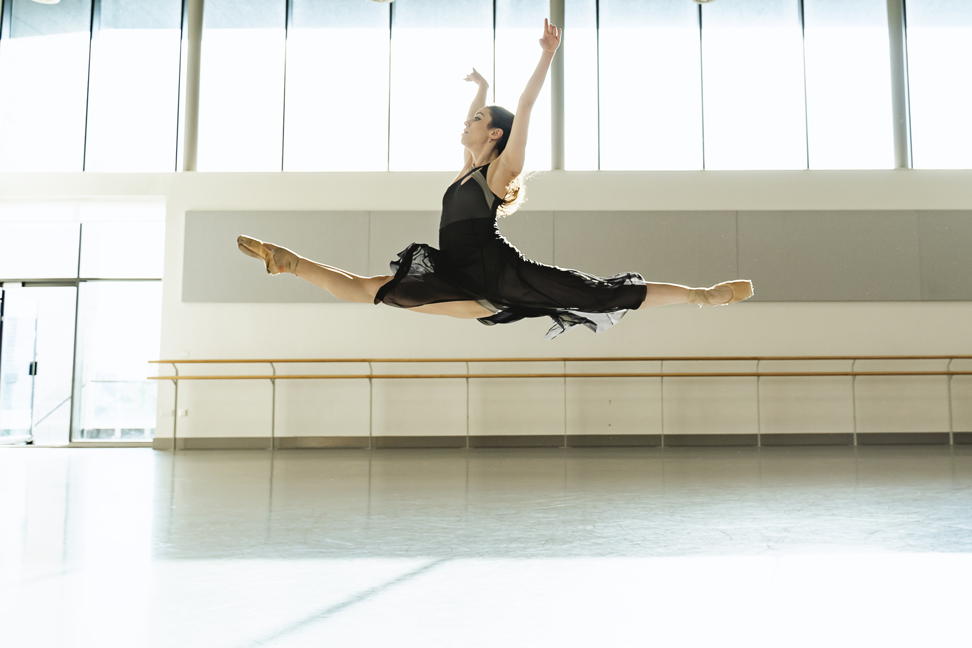 white female dancer in a split leap with arms up wearing a black skirt and leotard. In a dance studio. 