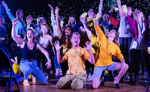 group of people wearing colour clothes on stage waving arms in the air with confetti in the air