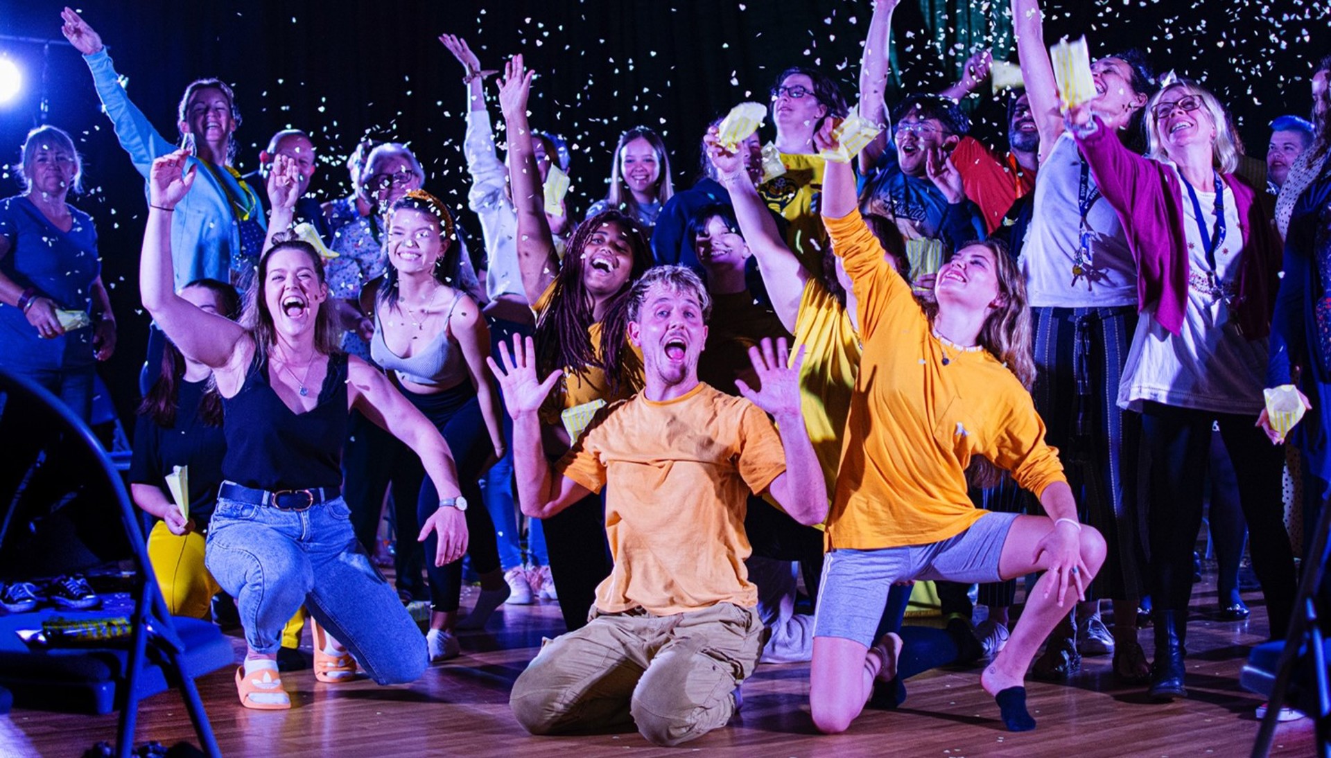 group of people wearing colour clothes on stage waving arms in the air with confetti in the air