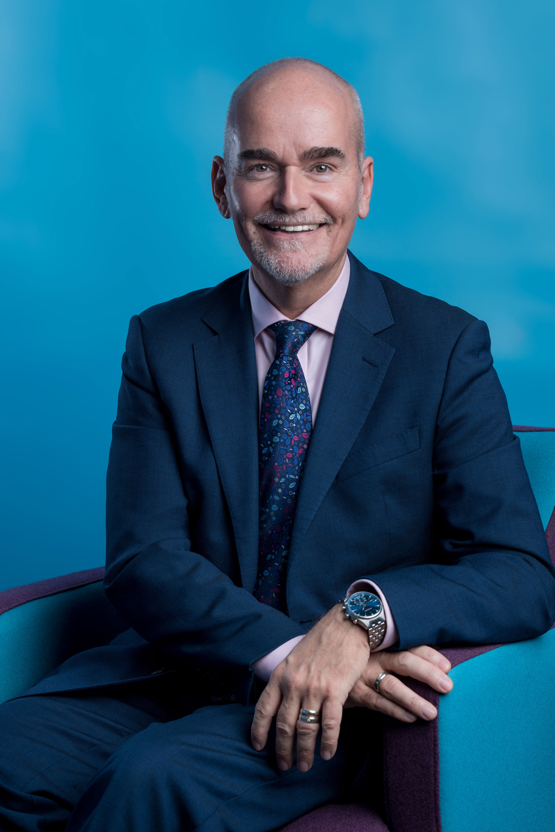 Headshot of David Blackburn. White man with shaved head smiling at the camera wearing and blue suit and tie