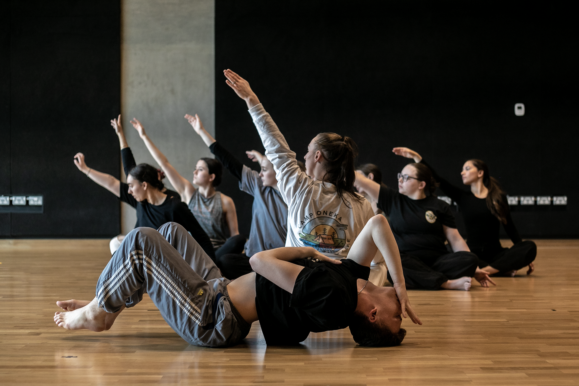 7 female dancers sitting crossed legged with one arm in the air. one male dancer infront laying on the floor with knees up arched back and hand infront of face. In a dance stucio with wooden floors and black walls 