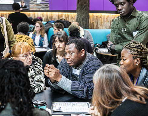 Global majority male in the centre of the image sitting at a table holding his hands together with a group of people listening to him of mixed ages, genders and ethnicities.  