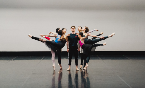 six females on point in an arabesque with leg high behind all holding onto centre male who is standing looking at the camera. In a dance studio 