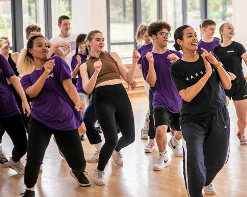 Female global majority dancer with curly hair tied back teaching a hip hop class. Students copying the lean back movement behind all wearing purple t-shirts in brightly lit dance studio