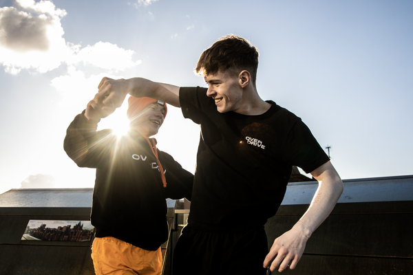 two young male dancers dancing together holding hands with lens flare inbetween them. wearing black and orange with one dancer in a beanie hat. infront of cloudy sky. 