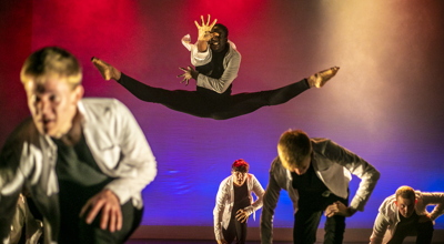 male dancers on U.dance stage wearing white shirts and black trousers. Four white young males crouched down one male black dancer jumpinging the box splits leaning forward with one hand stretched out. Lighting pink blue and red spotlights