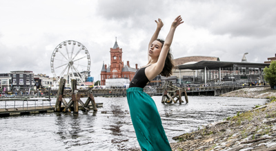 dancer with arched back and arms above head infront of Cardiff bay skyline. Young white female wearing black leotard and green silk trousers